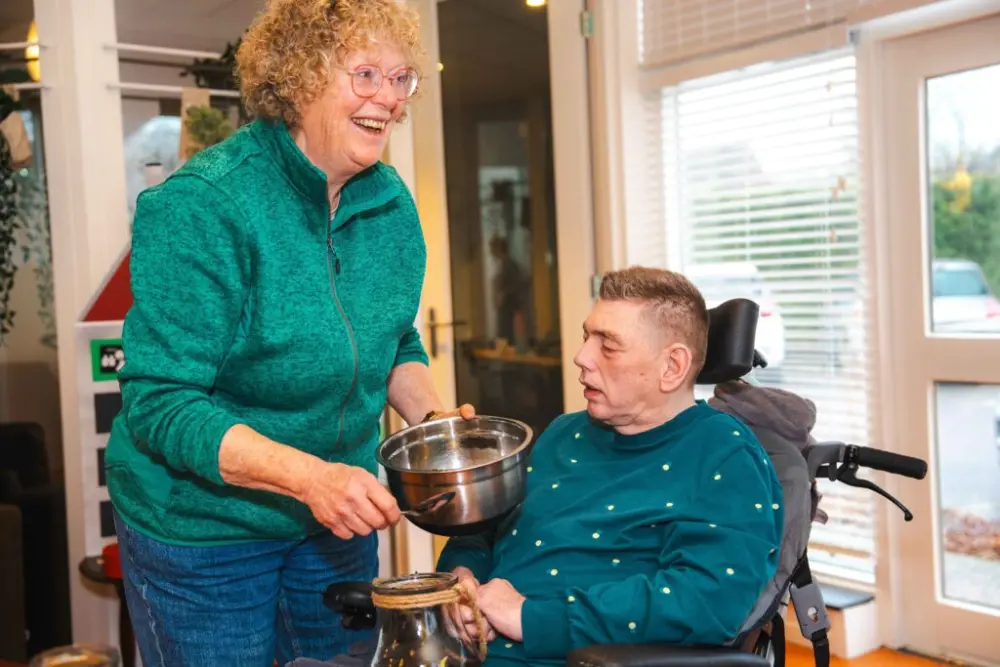 Een begeleider roert in een pan terwijl een bewoner in een rolstoel toekijkt in de keuken