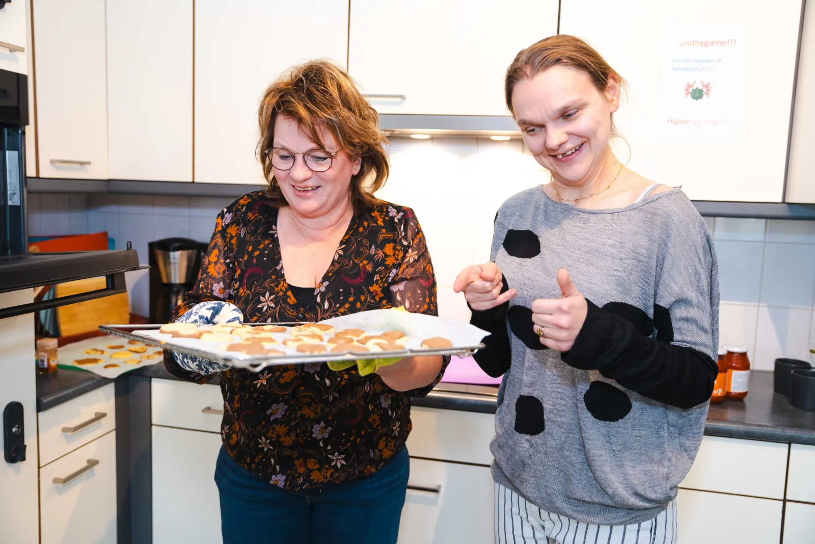 Een begeleider en een bewoner presenteren samen lachend een bakplaat met versgebakken koekjes in de woonkeuken
